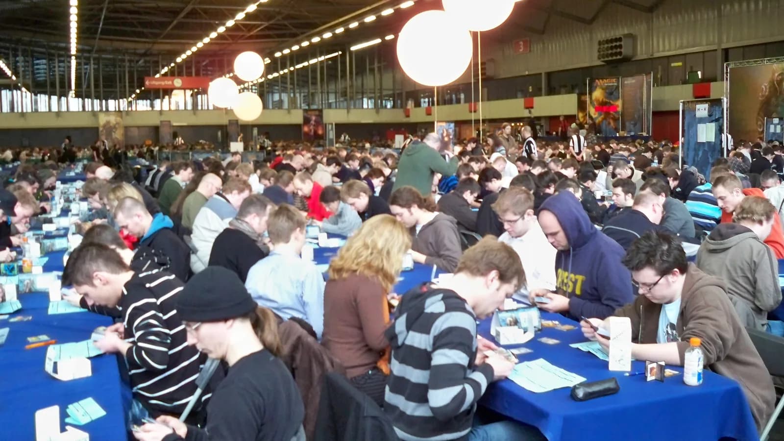 Salle de tournoi Magic the Gathering avec des centaines de joueurs autour de tables, kakémonos Magic en arrière-plan