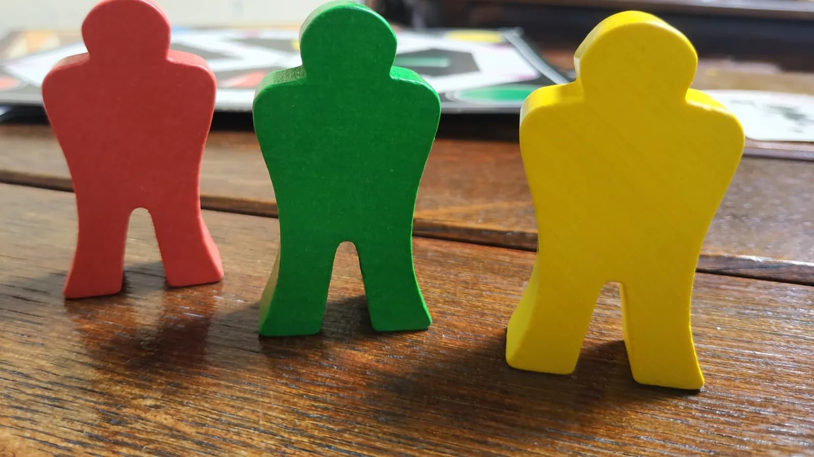 Trois meeples en bois colorés (rouge, vert, jaune) posés sur une table en bois devant un plateau de jeu