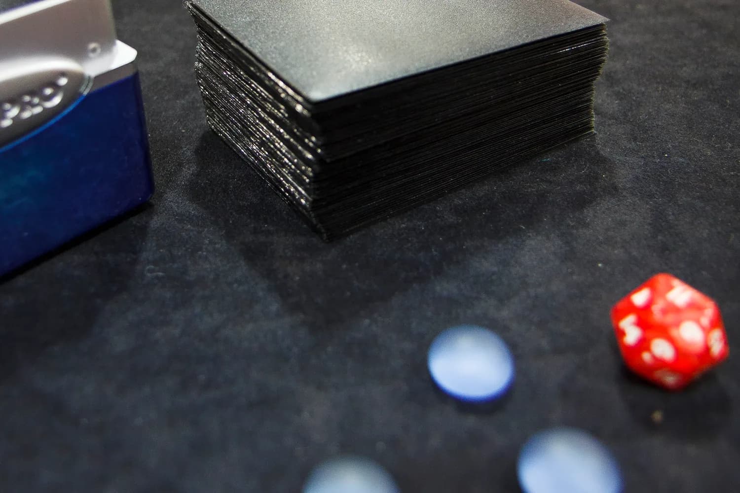 Deck de Magic the Gathering sous pochettes noires, boîte de rangement, jetons bleus et dé rouge à vingt faces posés sur un tapis de tournoi