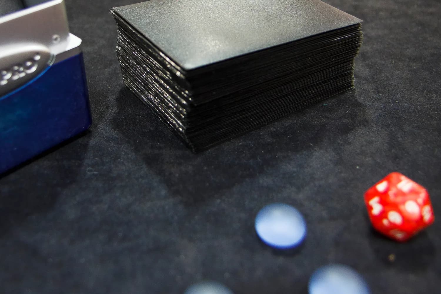 Deck de cartes Magic the Gathering sous pochettes noires, avec un dé rouge et des jetons bleus posés sur une table de tournoi
