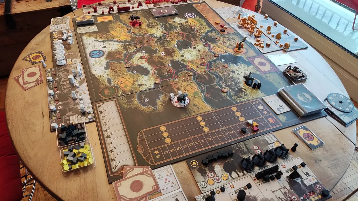 Plateau de Scythe mis en place sur une table : carte hexagonale, plateaux joueurs, figurines de mechs et jetons ressources